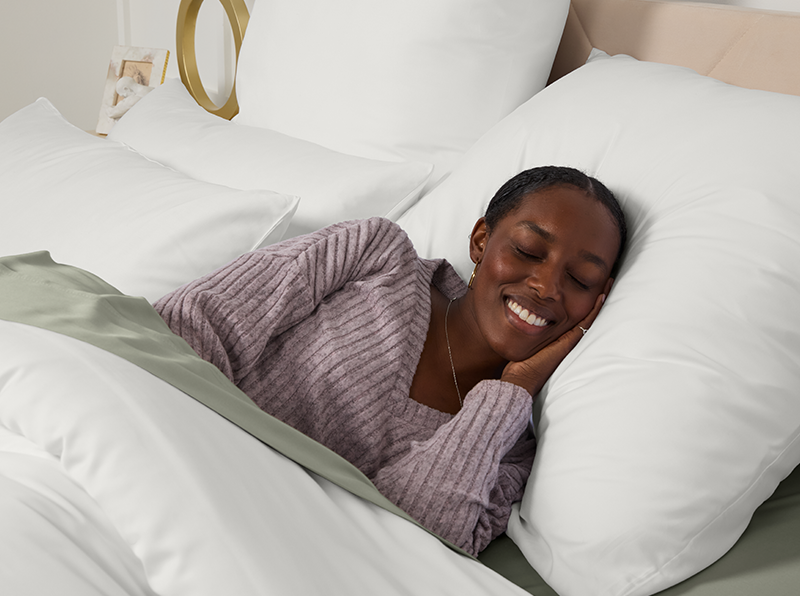 Woman peacefully sleeping in bed with white and sage green sheets, wearing a lavender sweater, smiling in comfort.