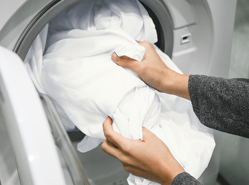 Hands loading soft white sheets into a washing machine, highlighting easy care and machine washability.