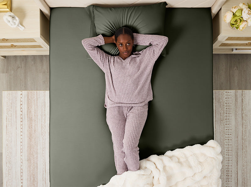 Woman lying on an olive green fitted sheet set, hands behind head, in cozy lavender pajamas on a neatly made bed.