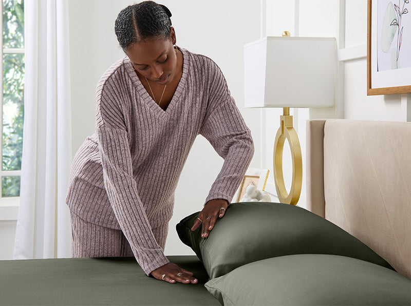 Woman in lavender loungewear smoothing olive green bed sheets on a neatly made bed in a bright, modern bedroom.